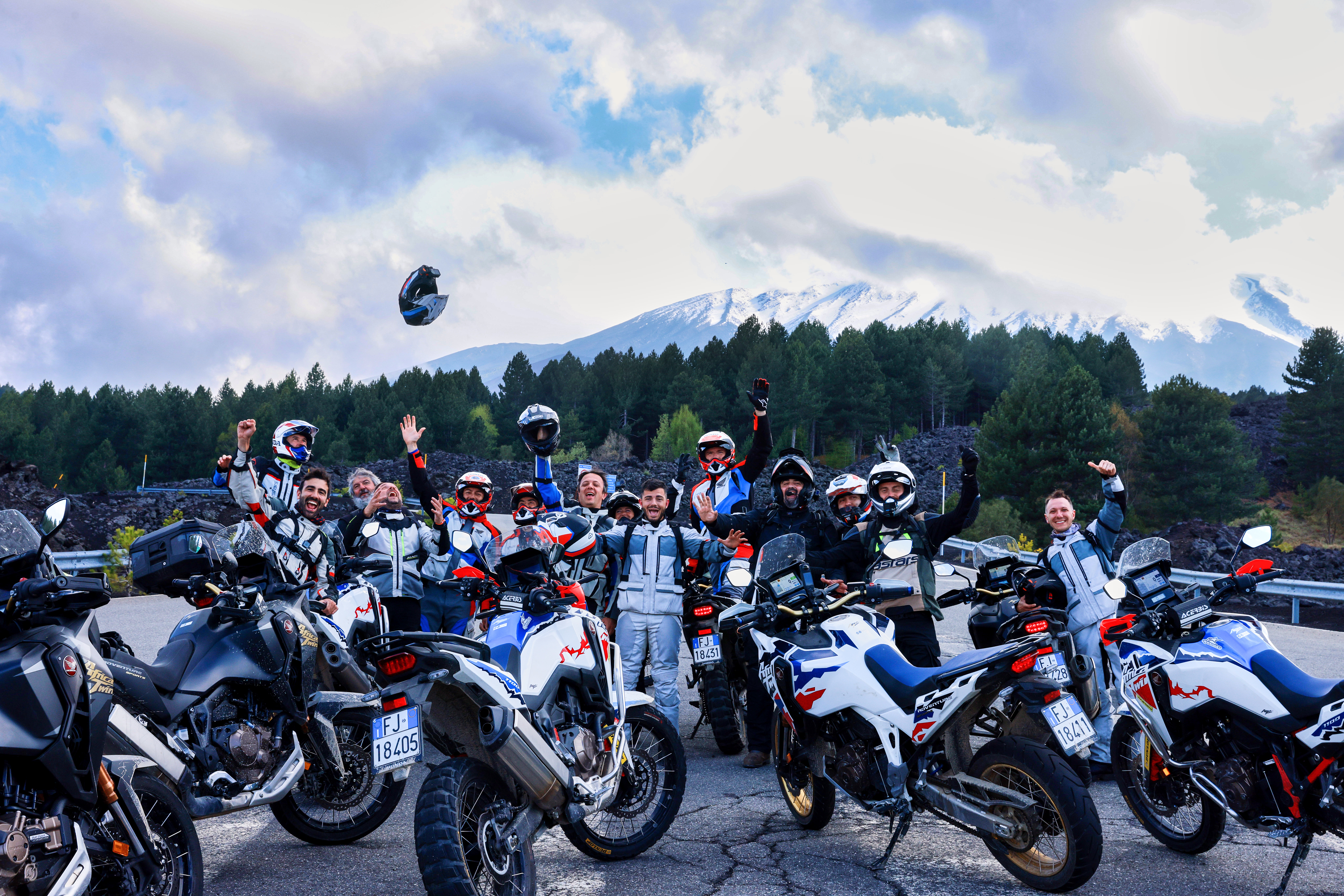 Un gruppo di motociclisti entusiasti, vestiti con tute da moto, che celebrano alzando i caschi in aria, con montagne innevate sullo sfondo.